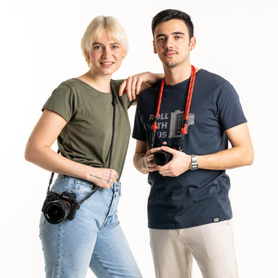Two photographers standing side-by-side one with an adjustable rope strap around their neck in red with a Leica Camera one with a Sony camera on their hip attached with a another strap 