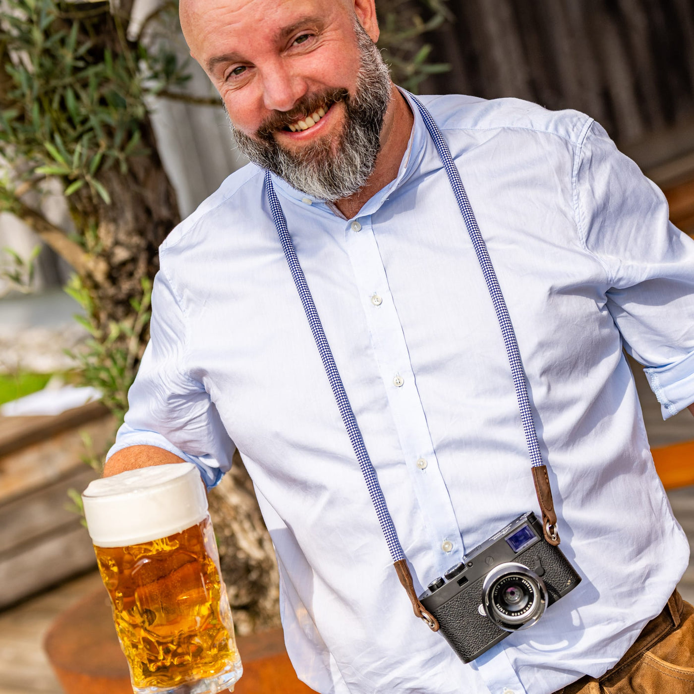 Man holding a glass of beer with a leica camera on the oktoberfest rope camera strap around his neck, standing outdoors. 