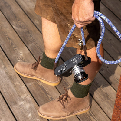 Person wearing brown leather shoes and green socks, holding a leica camera with the oktoberfest rope camera strap on a wooden floor. 