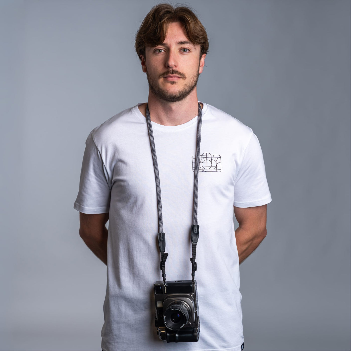 Photographer against dark gray background wearing a white t-shirt and the cooph rope camera strap with a vintage camera attached to it 