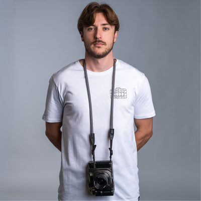 Photographer against dark gray background wearing a white t-shirt and the cooph rope camera strap with a vintage camera attached to it 