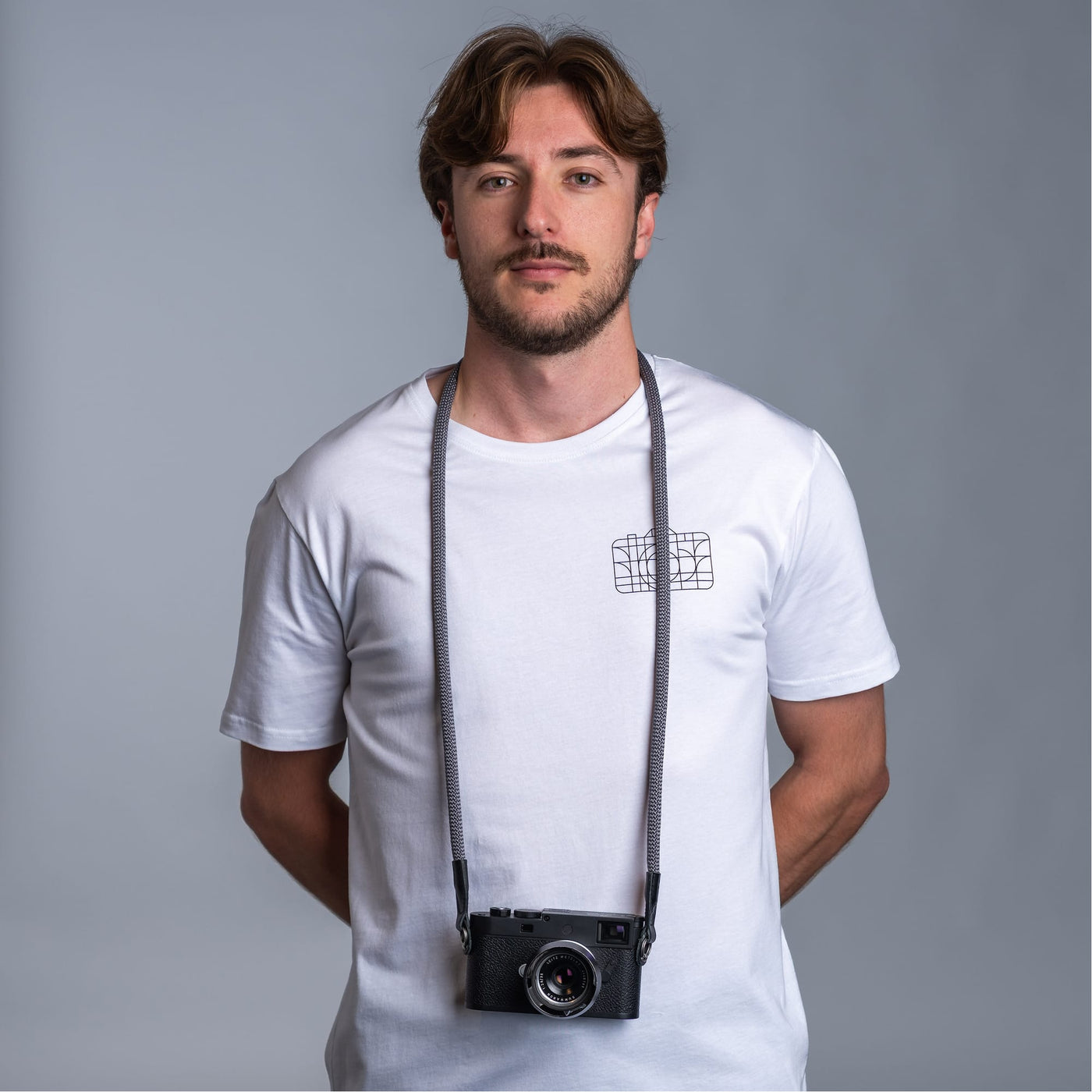 Photographer against dark gray background wearing a white t-shirt and the cooph rope camera strap with a leica camera attached to it 