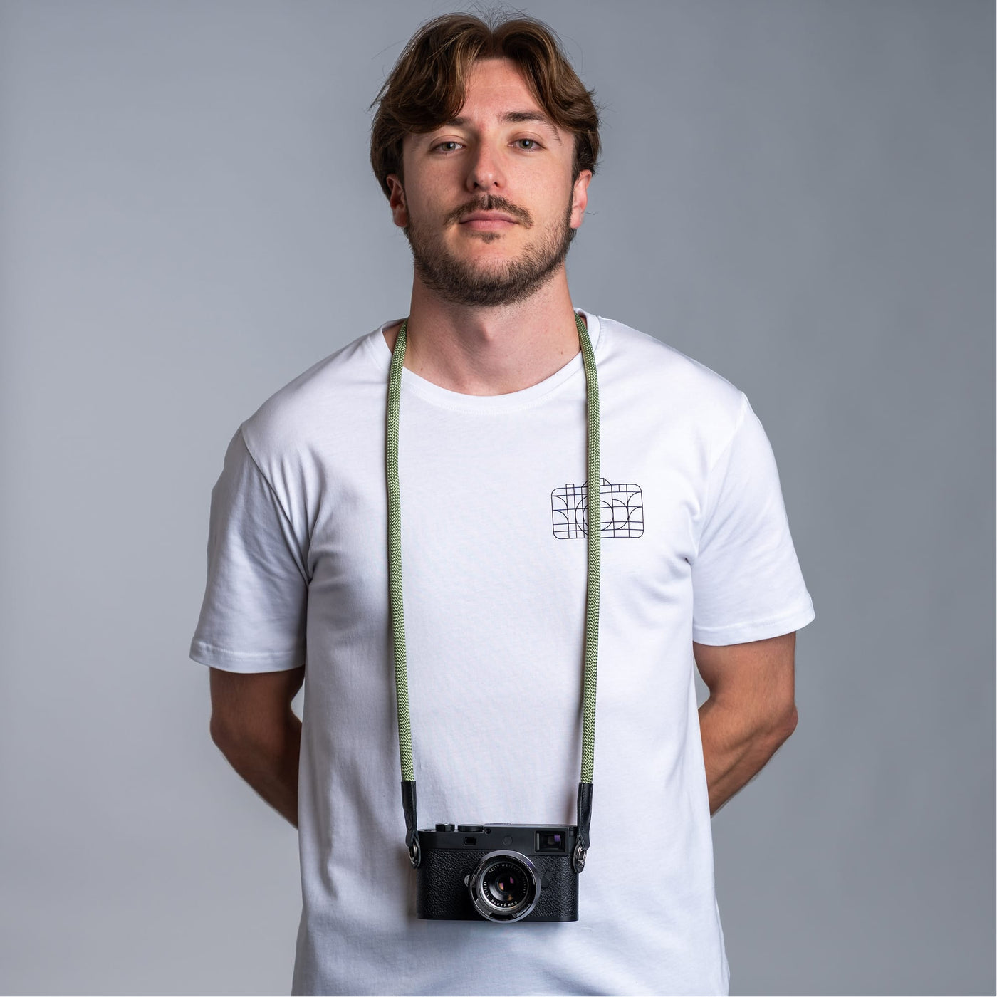 Photographer against dark gray background wearing a white t-shirt and the cooph rope camera strap with a leica camera attached to it 