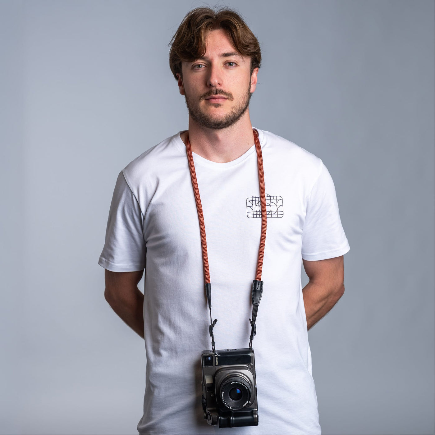 Photographer against dark gray background wearing a white t-shirt and the cooph rope camera strap with a vintage camera attached to it 