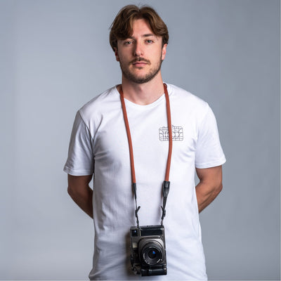 Photographer against dark gray background wearing a white t-shirt and the cooph rope camera strap with a vintage camera attached to it 