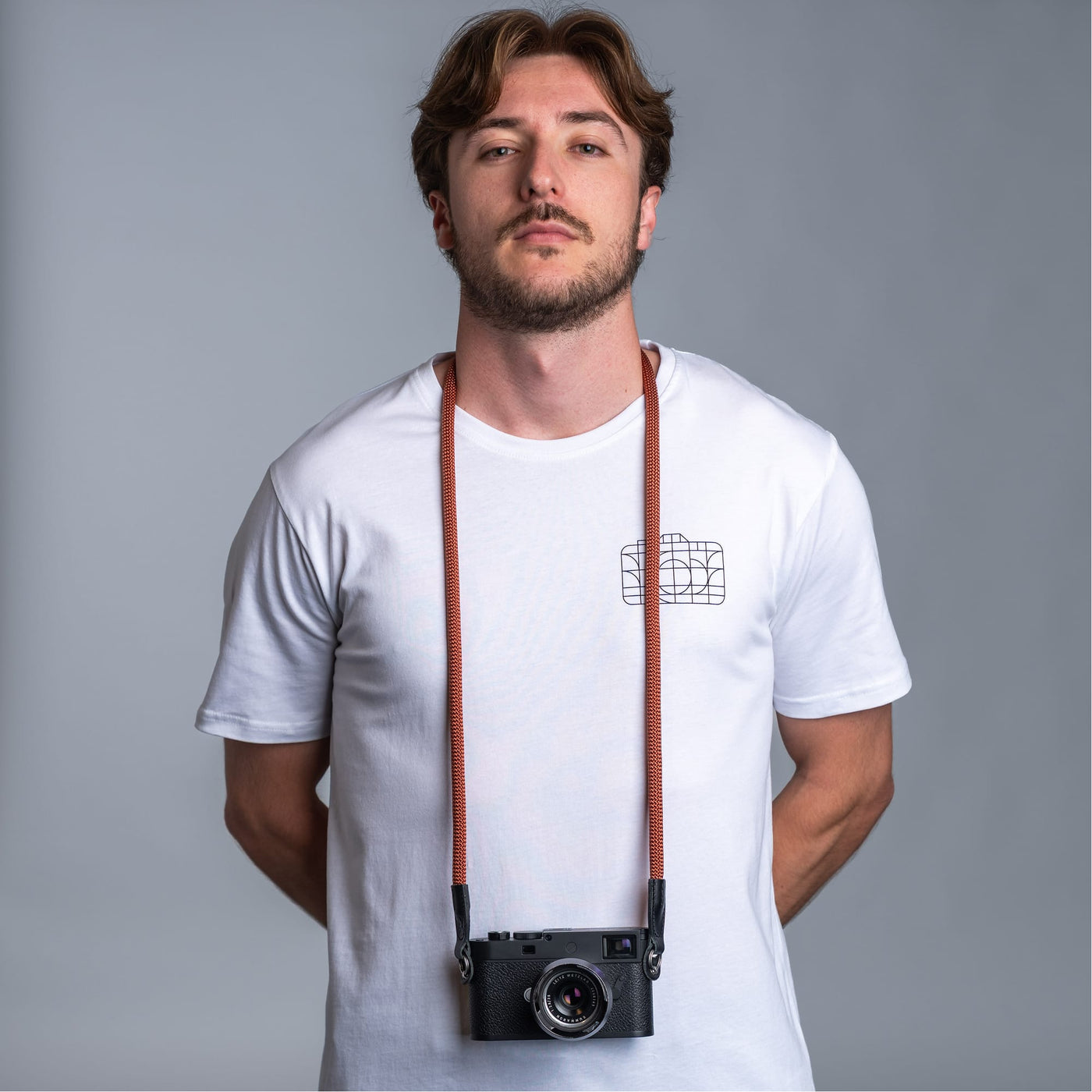 Photographer against dark gray background wearing a white t-shirt and the cooph rope camera strap with a leica camera attached to it 